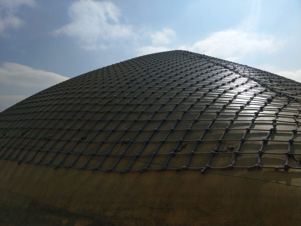 Foto von einem Teil einer großen gelben Hüpfpyramide mit einem Netz aus Kletterseilen drüber. Darüber ein wolkiger Himmel.