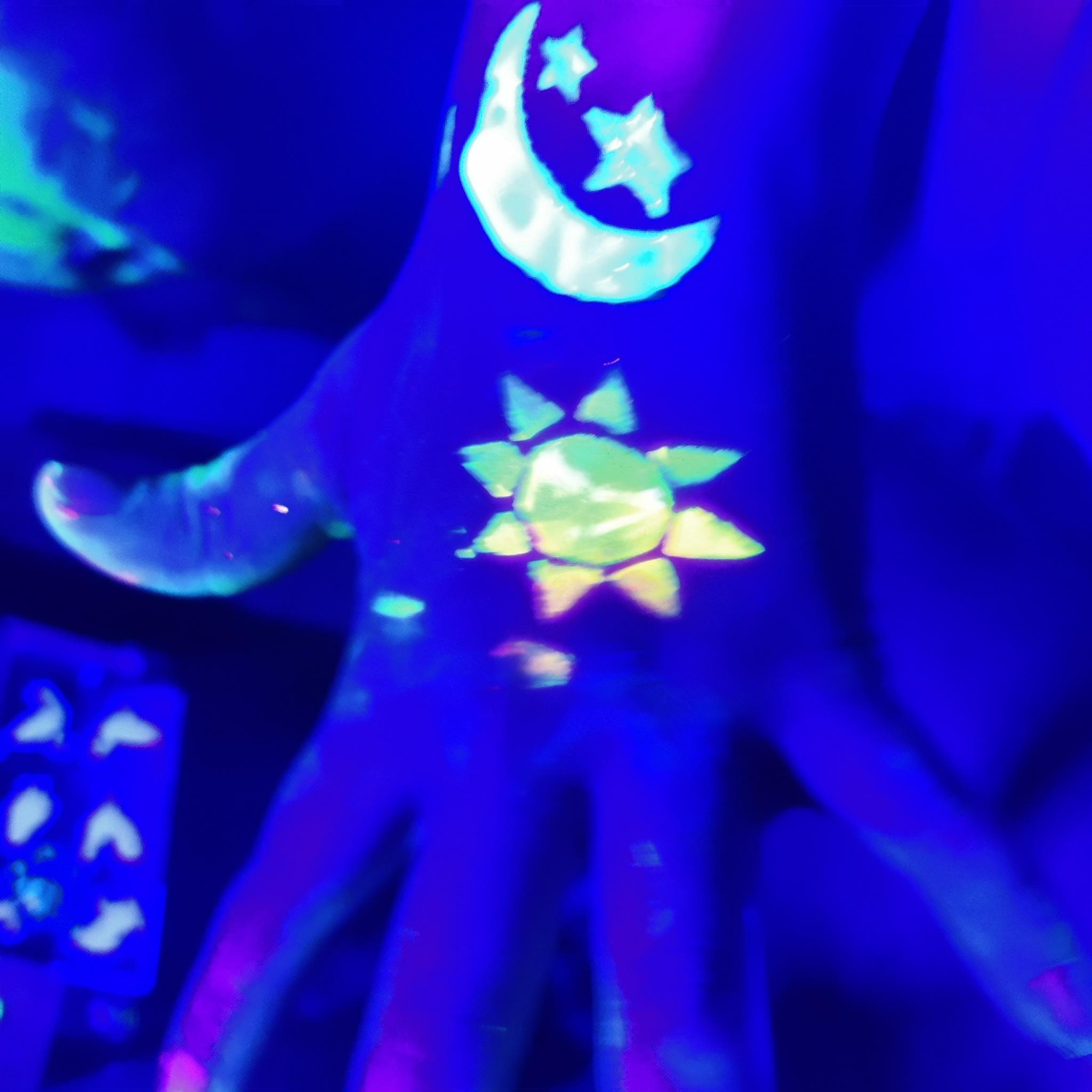 Rücken einer Hand mit fluroszierender Farbe. Eine gelbe Sonne auf dem Handrücken, eine Mondsichel und zwei Sterne auf dem Handgelenk. Im Hintergrund vereinzelt fluorszierende Farbflecken und Formen. Das Foto ist mit Schwarz-Licht aufgenommen, so dass die fluroszierenden Farben hervorstechen und der Rest diffus und dunkel ist.